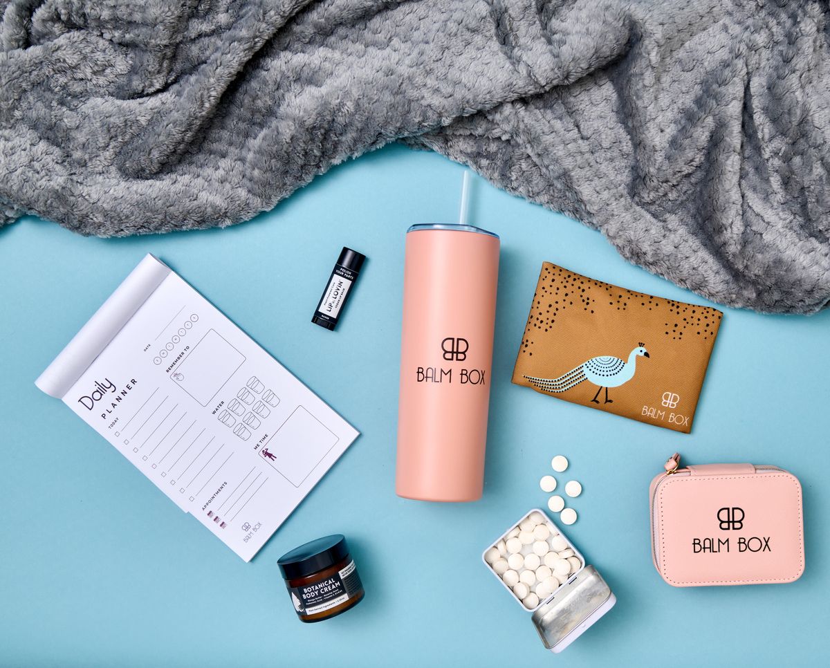 Flat lay of Balm Box items: gray throw blanket, pink tumbler, daily planner, lip balm, body cream, ginger mints, pink travel pill case, and an ice pack with a peacock design.